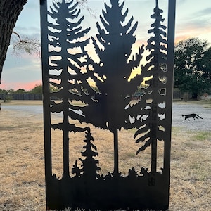 May include: A black metal silhouette of a forest scene with tall pine trees and a smaller tree in the foreground. The silhouette is cut out of a metal sheet and is mounted on a wooden frame.