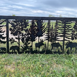 May include: Black metal silhouette of a forest scene with trees, moose, and bears. The silhouette is cut out of a metal sheet and mounted on a wooden post.