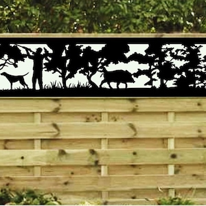 May include: A black metal fence panel with a silhouette design of hunters, a dog, and a wild boar in a forest setting. The panel is mounted on a wooden fence.