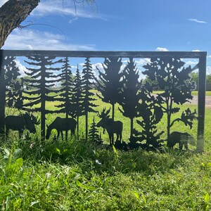 May include: A black metal fence panel with a cut-out design of trees and moose. The fence panel is in front of a grassy field with a blue sky and white clouds in the background.