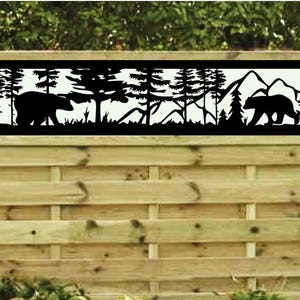 May include: Black metal fence panel featuring a forest scene with two bears, trees, and mountains. The panel is mounted on a wooden fence. The design is a silhouette against a white background.