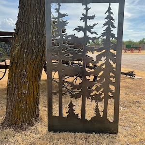 May include: A metal silhouette of a forest scene with multiple pine trees cut out of the metal. The metal is a dark gray color and the background is a blurry image of a grassy field.