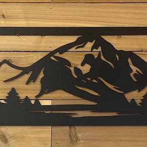 May include: Black metal wall art featuring a mountain range and forest silhouette within a rectangular frame. The artwork depicts detailed trees and peaks, creating a nature-inspired scene. The piece is designed to be mounted on a wall.