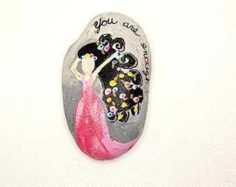 May include: Painted rock with a woman in a pink dress and black hair with flowers. The text "you are enough" is written on the rock.