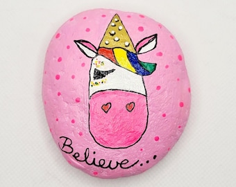 May include: A pink painted rock with white polka dots. The rock has a painted unicorn with a rainbow mane and a gold crown. The unicorn has two pink hearts on its face. The rock has the word "Believe..." written on it.