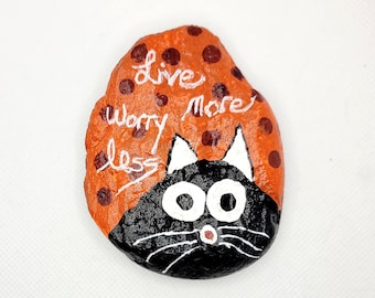 May include: Painted rock with a black cat face and the words "Live more worry less" in white on an orange background with brown polka dots.
