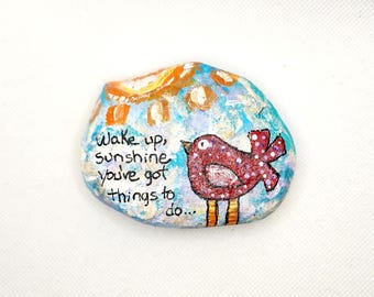 May include: Painted rock with a blue, white, and orange background. A red bird with white polka dots and yellow legs is painted on the rock. The text "Wake up, sunshine you've got things to do..." is written on the rock.