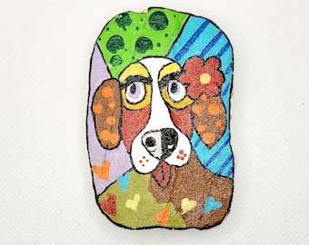 May include: A painted rock with a dog face. The dog has brown, white, and red fur. The rock is painted in a colorful patchwork style with green, blue, purple, and orange.