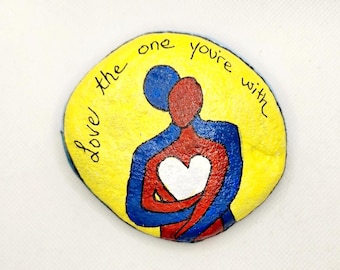 May include: Painted rock with a yellow background, a blue and red silhouette of two people embracing, and a white heart in the center. The text "love the one you're with" is written around the figures.