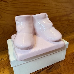 Könnte beinhalten: Eine hellrosa Keramik-Babyschuh-Skulptur. Die Schuhe sind auf einer passenden rosa rechteckigen Basis platziert, die sich auf einer weißen Box befindet. Die Skulptur ist ein dekorativer Artikel, möglicherweise für ein Kinderzimmer oder eine Baby-Themen-Dekoration.