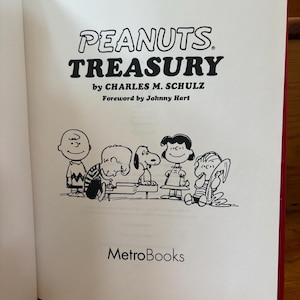 May include: Open book with the title "PEANUTS TREASURY" by Charles M. Schulz. The book's interior features a black and white illustration of Peanuts characters, including Charlie Brown, Snoopy, and Lucy. The MetroBooks logo is at the bottom.