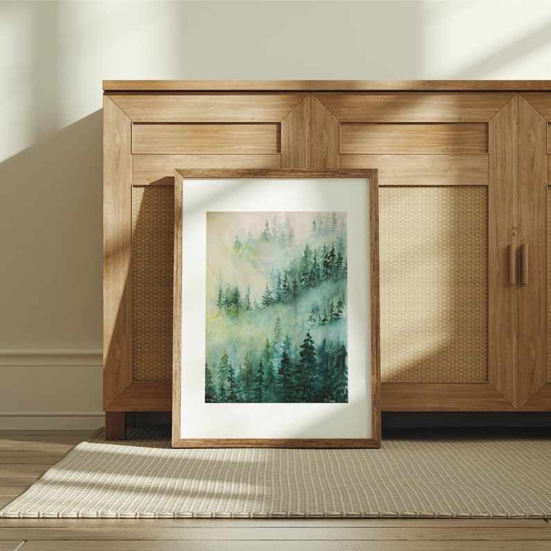 May include: A framed watercolor painting of a misty forest scene with green trees and a light blue sky. The painting is in a simple brown frame and is hanging on a wall.