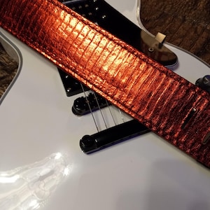 May include: A red guitar strap with a textured, reptile-like pattern is draped over a white electric guitar. The strap's color is a vibrant, metallic red, and the guitar has black hardware.