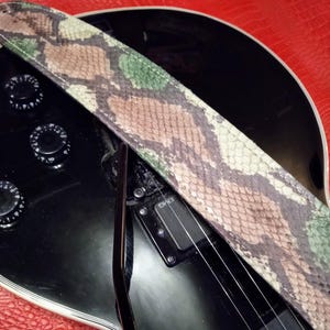 May include: A close-up of a black electric guitar with a green, brown, and white snake skin guitar strap. The guitar has a black pickguard and a single pickup.