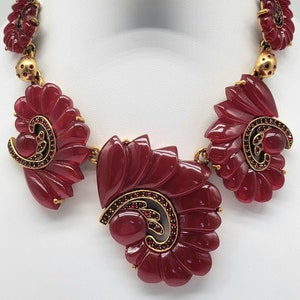 May include: A statement necklace featuring large, burgundy-colored, fan-shaped pendants. The necklace has gold-toned accents and is adorned with small red rhinestones. The design is displayed on a white jewelry stand.