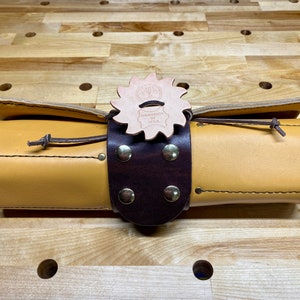 May include: A rolled-up leather tool roll with a brown leather strap and brass rivets. The strap is secured with a leather buckle with a decorative, star-shaped leather tag that reads "Handmade in Texas".