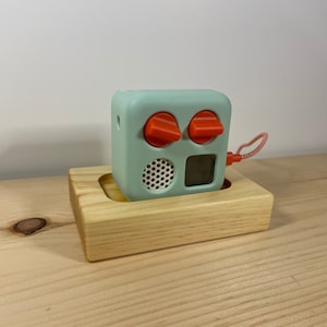 May include: A mint green cube-shaped audio player with two orange knobs and a small screen, resting in a light wood holder. The player has a speaker and a red and white cord. The holder is made of light-colored wood.