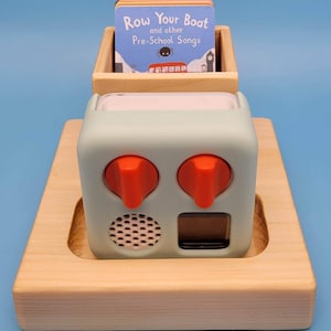 May include: A light green toy with two orange knobs, a speaker, and a small screen, set on a wooden tray. A small wooden box on top holds a book titled "Row Your Boat and other Pre-School Songs".