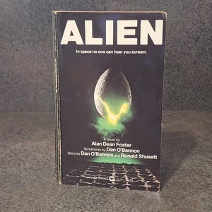 May include: A well-worn paperback book titled "ALIEN" with a dark cover. The cover art shows an alien egg with green mist and the text "In space no one can hear you scream". It is a novel by Alan Dean Foster.