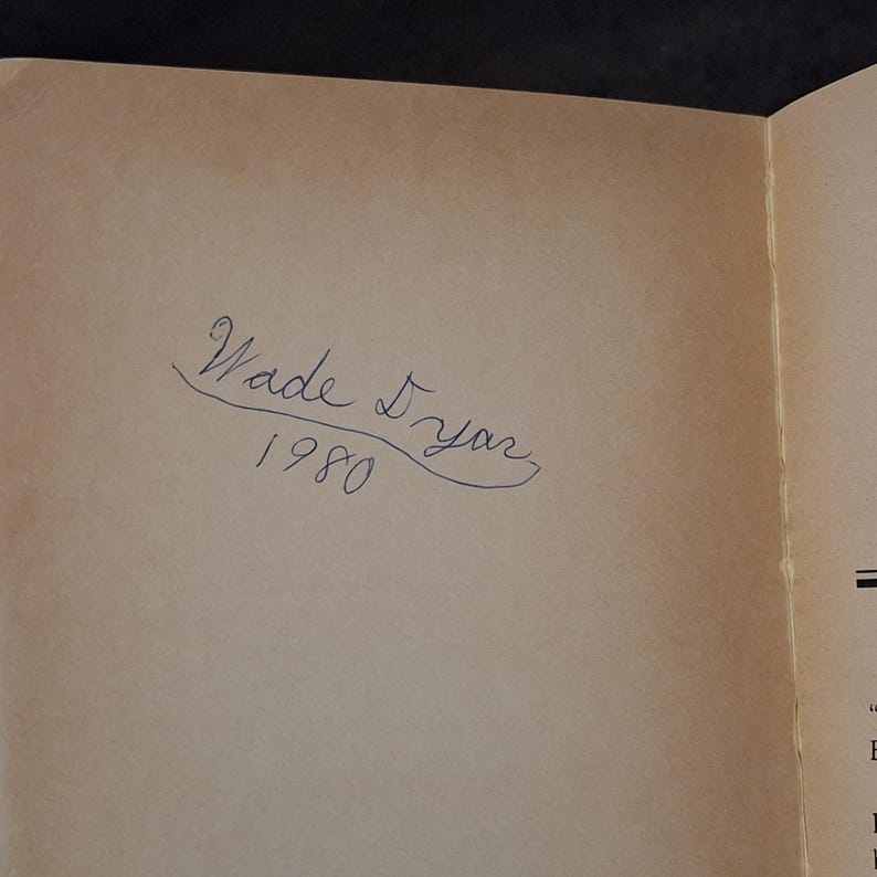 May include: An open book page with the handwritten inscription "Wade Dyar 1980" in blue ink. The paper has a light brown color.
