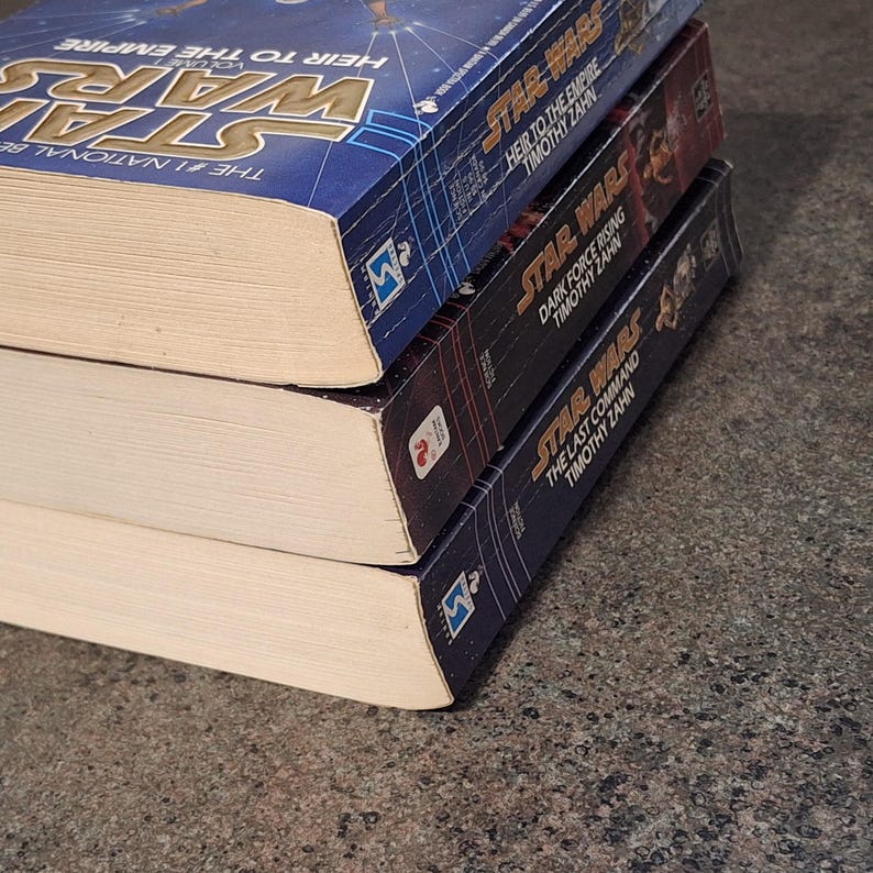 Op de afbeelding: Drie Star Wars-romans op elkaar gestapeld. Het bovenste boek is blauw met de titel "Heir to the Empire". Het middelste boek is zwart met de titel "Dark Force Rising". Het onderste boek is donkerblauw met de titel "The Last Command".