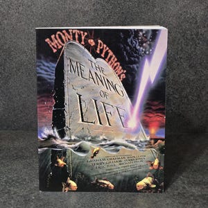 Op de afbeelding: Een boek getiteld "Monty Python's The Meaning of Life" met een dramatische omslagillustratie. De omslag toont een stenen tablet die uit het water oprijst, met de titel erop. Een bliksemschicht schiet door een stormachtige lucht.