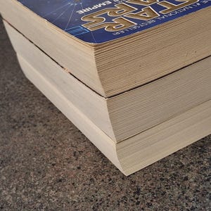 Op de afbeelding: Close-up van drie boeken die op elkaar zijn gestapeld. Het bovenste boek heeft een blauwe kaft met de woorden "STAR WARS" en "THE EMPIRE" in wit en geel. De pagina's van het boek zijn beige.