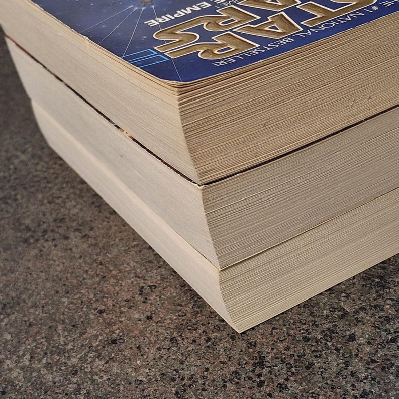 Op de afbeelding: Close-up van drie boeken die op elkaar zijn gestapeld. Het bovenste boek heeft een blauwe kaft met de woorden "STAR WARS" en "THE EMPIRE" in wit en geel. De pagina's van het boek zijn beige.