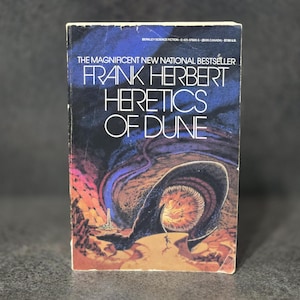 May include: A paperback book titled "Heretics of Dune" by Frank Herbert. The cover features a dark blue background with a large, dark creature and a small figure in a desert landscape. The title is in white, with the author's name above.