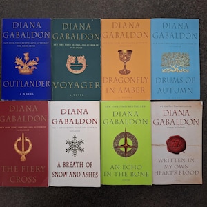 May include: A collection of eight books by Diana Gabaldon arranged in two rows. The titles include Outlander, Voyager, Dragonfly in Amber, Drums of Autumn, The Fiery Cross, A Breath of Snow and Ashes, An Echo in the Bone, and Written in My Own Heart's Blood.