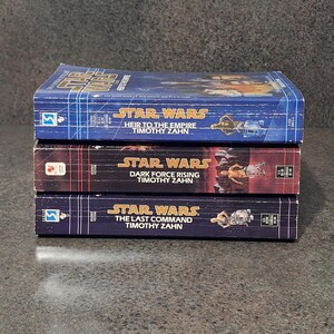 Op de afbeelding: Drie Star Wars-romans van Timothy Zahn zijn op elkaar gestapeld. De boeken heten "Heir to the Empire", "Dark Force Rising" en "The Last Command". Elk boek heeft het Star Wars-logo en illustraties van personages. De boeken liggen op een donkere ondergrond.