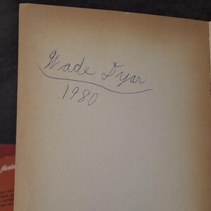 May include: A page from a book with the handwritten inscription "Wade Dyer 1980" in blue ink. The paper has a light brown, aged appearance.