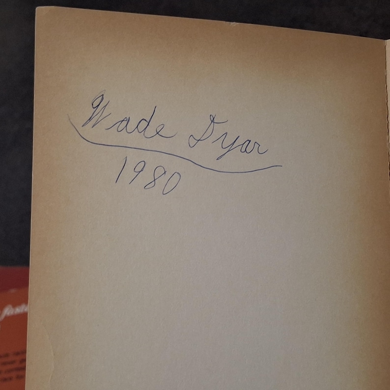 May include: A page from a book with the handwritten inscription "Wade Dyer 1980" in blue ink. The paper has a light brown, aged appearance.