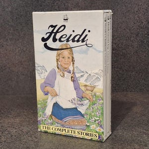 Könnte beinhalten: Ein gebundenes Buch mit dem Titel "Heidi", das ein junges Mädchen in traditioneller Kleidung vor einer Berg- und Blumenlandschaft zeigt. Auf dem Cover steht "THE COMPLETE STORIES".