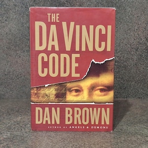 Op de afbeelding: Een hardcover boek getiteld The Da Vinci Code van Dan Brown. De rode kaft bevat grote beige tekst en een gescheurd horizontaal gedeelte dat een close-up van de ogen van de Mona Lisa en handgeschreven tekst onthult. Het andere werk staat onderaan.