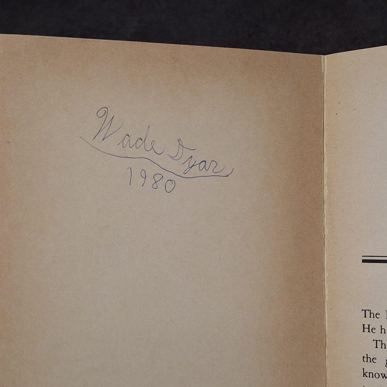 May include: An open book with a light brown page. The handwritten text "Wade Syar 1980" is in black ink. The page is slightly aged, with a visible crease.