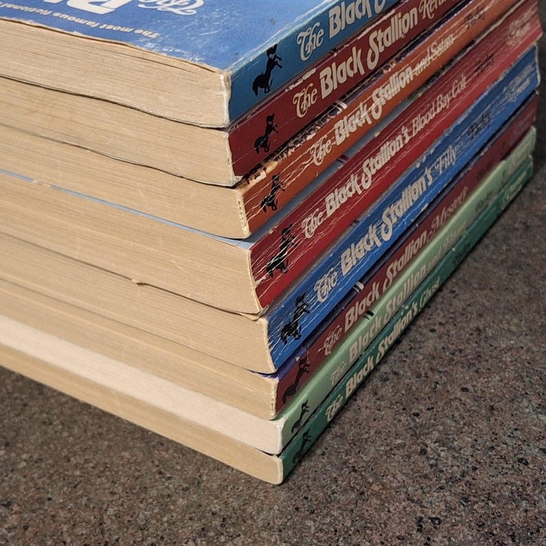May include: A stack of vintage paperback books featuring "The Black Stallion" series. The books have colorful covers in blue, red, and green, with titles and a black horse silhouette. The pages are aged, showing signs of wear.