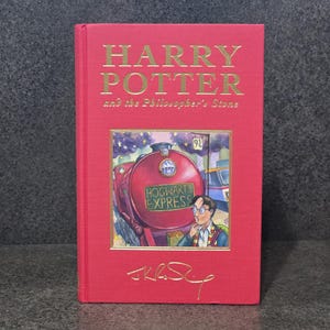 May include: A red hardback book titled "Harry Potter and the Philosopher's Stone" with gold lettering. The cover shows the Hogwarts Express train and a young man. The author's signature is at the bottom.