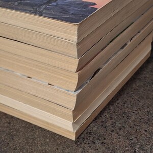 May include: A stack of vintage books with aged, yellowed pages. The covers show hints of illustrations and titles, with a focus on the worn edges and the texture of the paper. The books are stacked on a textured surface.