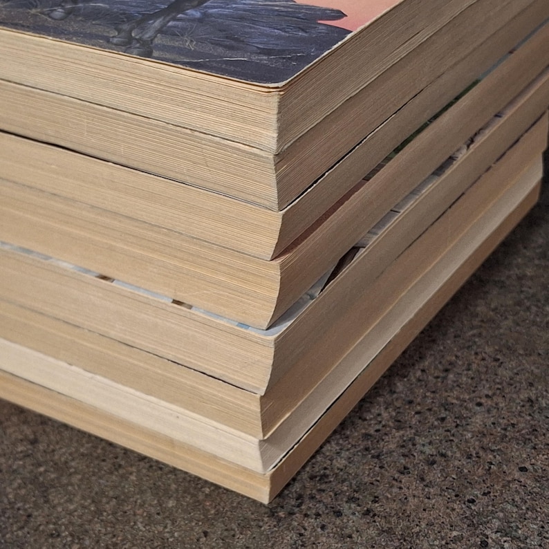 May include: A stack of vintage books with aged, yellowed pages. The covers show hints of illustrations and titles, with a focus on the worn edges and the texture of the paper. The books are stacked on a textured surface.