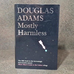 Könnte beinhalten: Ein dunkelblaues Hardcover-Buch mit dem Titel "DOUGLAS ADAMS Mostly Harmless" mit silberner Schrift. Das Cover zeigt einen weißen Pfeil, der auf einen blauen Punkt zeigt, mit dem Text "Das fünfte Buch der zunehmend unzutreffend benannten Hitch Hiker's Guide to the Galaxy-Trilogie."