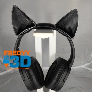 Puede incluir: Auriculares negros con orejas de gato unidas a la diadema. Los auriculares están en un soporte blanco. El texto "FREDYY 3D" es visible en la copa izquierda.