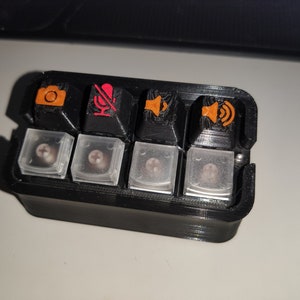 Könnte beinhalten: Ein schwarzes Plastikgehäuse mit sechs durchsichtigen Plastikknöpfen. Die Knöpfe haben orange und rote Symbole, darunter eine Kamera, ein Mikrofon und ein Lautsprecher.
