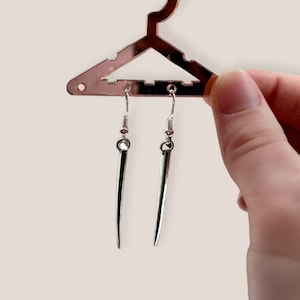 May include: A pair of silver-colored, pointed earrings hanging from a rose gold-colored hanger. The earrings have a simple, elongated design with a hook closure. The hanger is shaped like a clothing hanger.