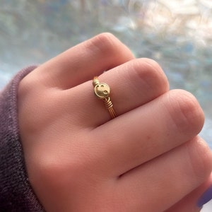 May include: A gold-colored wire ring featuring a small smiley face charm. The ring's band is made of multiple strands of gold wire, and the smiley face charm is centered on the ring. The ring is shown on a hand.