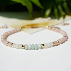 Könnte beinhalten: Zartes Perlenarmband mit Rosenquarz-, weißen und hellgrünen Edelsteinen. Das Armband hat goldfarbene Akzente und ist auf einer neutralfarbenen Kordel aufgefädelt. Es ist ein Schmuckstück.