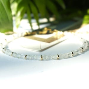 Op de afbeelding: Een verfijnde armband bestaande uit een enkele streng van kleine, gefacetteerde, doorschijnende witte maansteen kralen afgewisseld met piepkleine, gepolijste goudkleurige metalen tussenkralen, voorzien van een goudkleurige sluiting en een verstelketting.