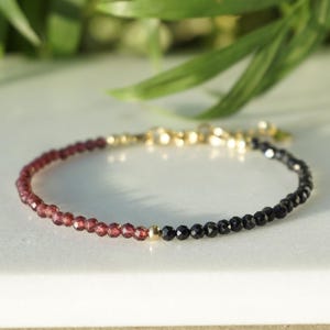 Könnte beinhalten: Ein zartes Armband mit einem Farbverlauf aus kleinen, facettierten Perlen. Das Armband wechselt von tiefem Rot zu Schwarz, mit einem goldfarbenen Verschluss und Akzenten. Der Schmuck wird auf einer weißen Oberfläche mit grünem Laub im Hintergrund präsentiert.