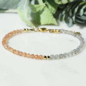 Op de afbeelding: Een delicate armband met een rij gefacetteerde oranje zonnesteenkralen aan de ene kant en gefacetteerde grijze labradorietkralen aan de andere kant, gescheiden door twee kleine goudkleurige metalen kralen en een goudkleurige karabijnsluiting.
