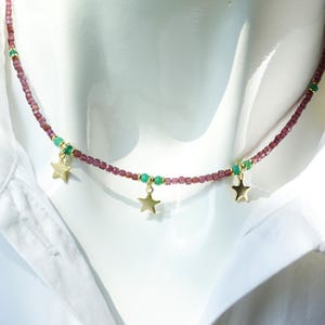 Puede incluir: Un delicado collar con una hebra de pequeñas cuentas rojo oscuro intercaladas con cuentas verdes. Los dijes de estrella dorados cuelgan del collar, creando un diseño celestial. El collar se exhibe sobre una superficie blanca.